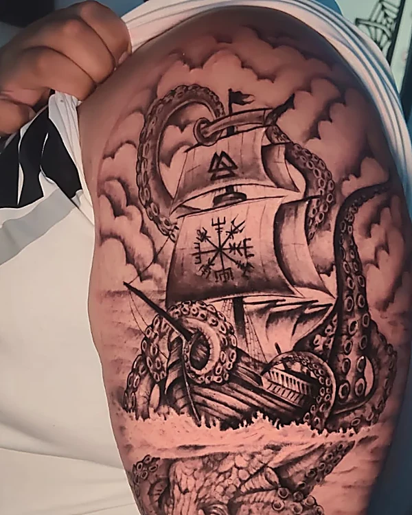 Kraken enroscado en un barco de vela con brújula vikinga, en negro y gris, tatuado en la parte alta del brazo