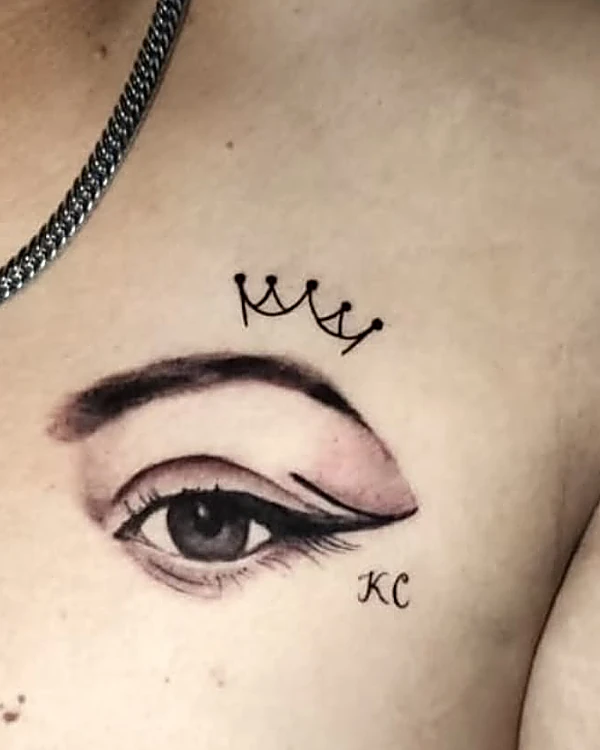 Ojo delineado en negro y gris con una corona pequeña y las iniciales KC, cerca de la clavícula